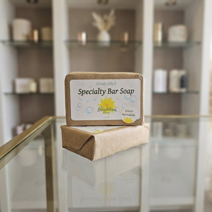 Photograph the 'Specialty Bar Soap' collection as standalone pieces on a pristine, reflective glass display in an upscale boutique setting. Utilize a combination of diffused daylight and rim lighting to emphasize the translucency and artisanal craftsmanshi