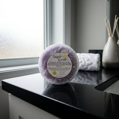 Display the 'Sugar Scrub' prominently in a luxurious bathroom scene, styled with modern minimalist decor. The jar is centered on a glossy black countertop, lit by diffused daylight through a frosted glass window, creating a bright, airy atmosphere. Use a D