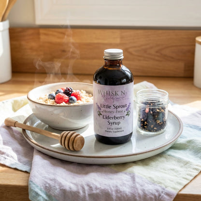 A lifestyle scene set in a brightly lit, cozy kitchen: the Little Sprout Elderberry Syrup bottle sits on a ceramic breakfast dish next to a steaming bowl of oatmeal topped with berries, a honey dipper, and a small jar of organic ingredients. High-key, diff
