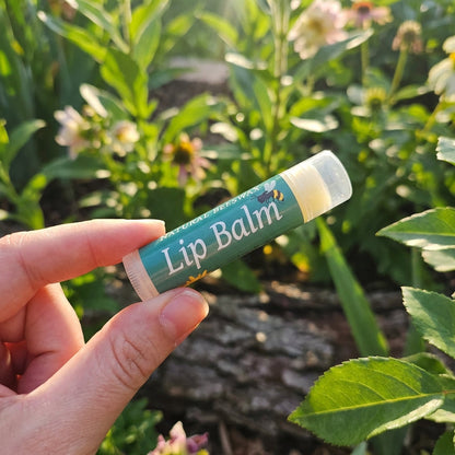 A lifestyle scene showcasing the 'Natural Bees Wax Lip Balm' held gently between fingers in a serene outdoor garden. Natural sunlight filtered through leaves creates dappled lighting, with a slight rim light emphasizing the product’s smooth, rounded form. 