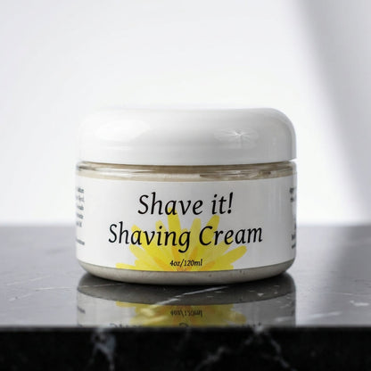 A high-resolution, close-up shot of the 'Shave it! Shaving Cream' container prominently positioned on a sleek, reflective black marble countertop, accented by softbox studio lighting that emphasizes the smooth, creamy texture of the cream and the bold, mod