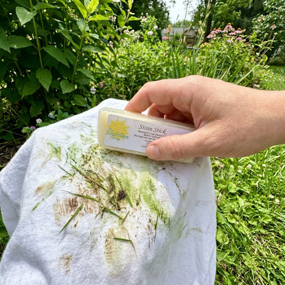 A dynamic composition showing the Stain Stick held in a hand above a stained white fabric, in an outdoor garden setting. Bright, natural diffused daylight from a high noon sun creates soft shadows and highlights the vibrant green of the grass and foliage i