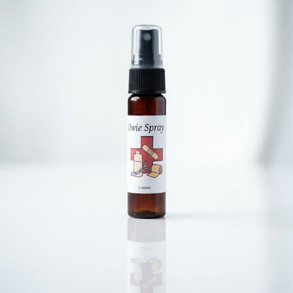 A close-up shot of Owie Spray standing upright on a sleek, reflective white surface in a professional studio setting. The lighting is softbox diffused daylight, emphasizing the clear, clean design of the spray bottle with detailed visibility of the label s