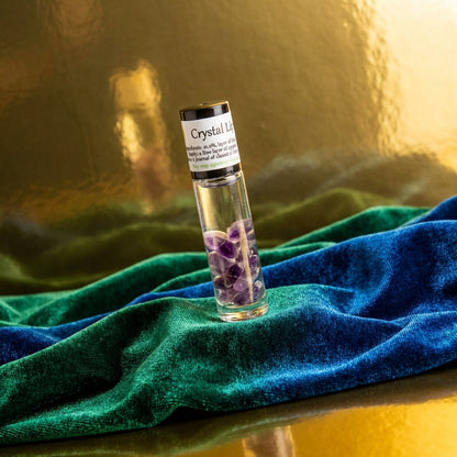 A high-fashion editorial style image with the Crystal Lip Gloss standing upright on a luxurious velvet cloth in rich jewel tones of emerald and sapphire. Studio HDR lighting creates dramatic highlights and deep shadows that enhance the crystal’s facets and