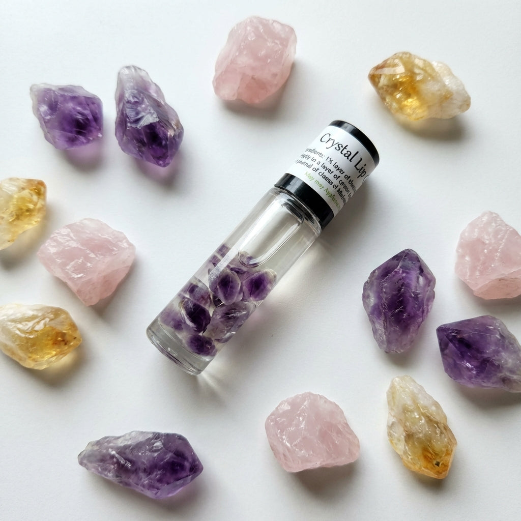 An artistic flat lay composition of the Crystal Lip Gloss tube surrounded by scattered crystals of amethyst, rose quartz, and citrine on a matte white surface. Soft diffused daylight from a nearby window creates gentle shadows and highlights the transparen