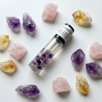 An artistic flat lay composition of the Crystal Lip Gloss tube surrounded by scattered crystals of amethyst, rose quartz, and citrine on a matte white surface. Soft diffused daylight from a nearby window creates gentle shadows and highlights the transparen